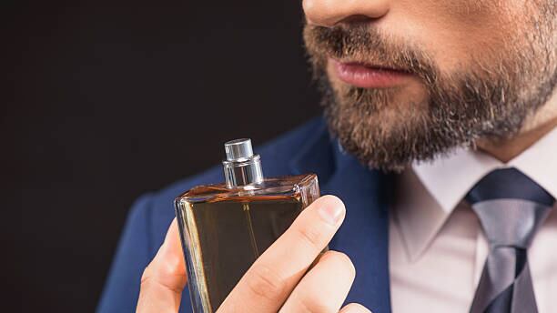 Homem de barba e e terno azul e segurando um frasco de perfume