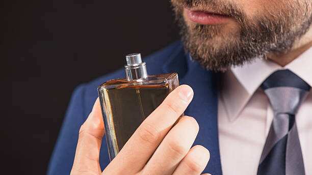 Homem de barba e e terno azul e segurando um frasco de perfume