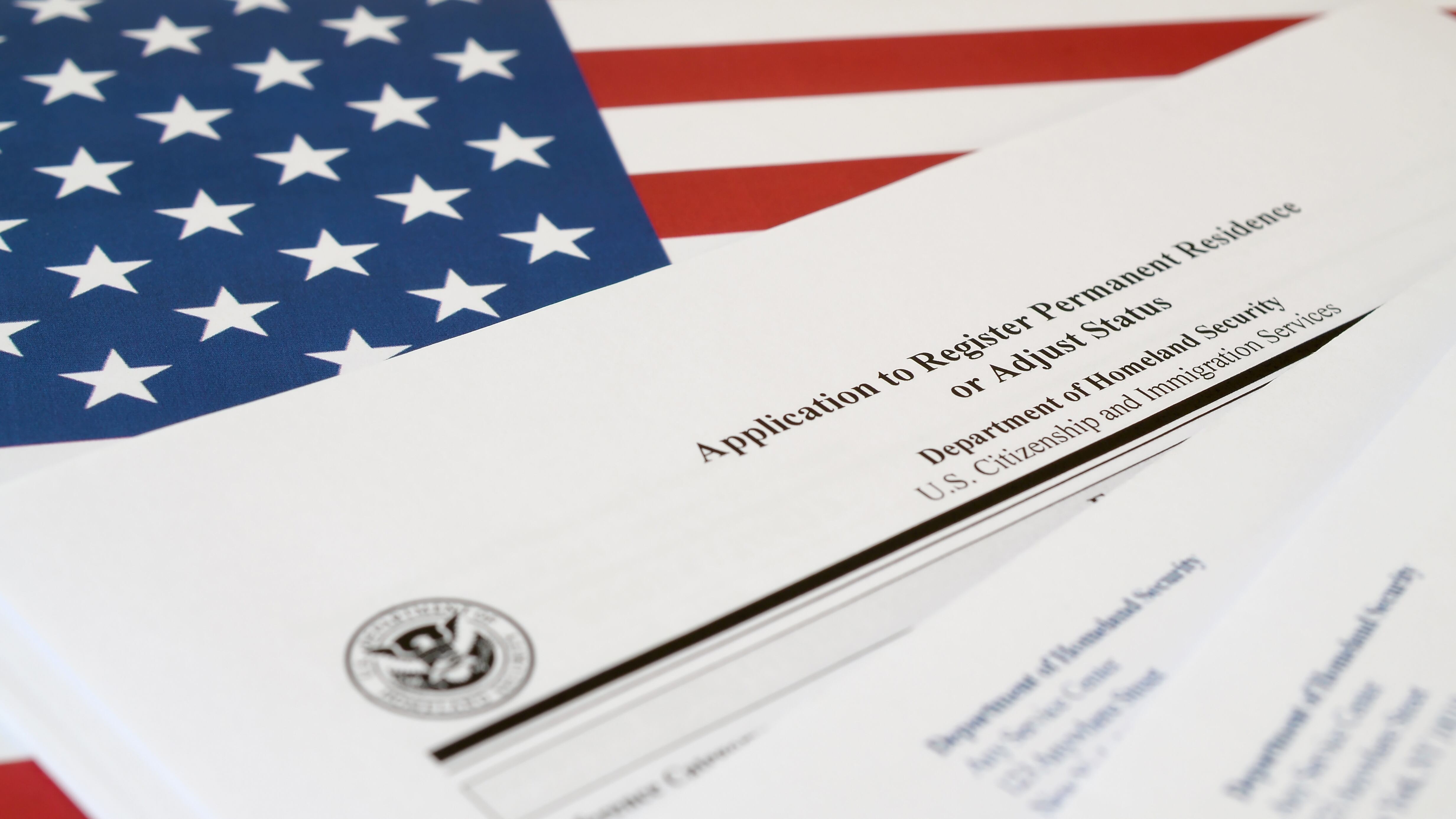 Formulario en blanco de la solicitud I-485 para registrar la residencia permanente o ajustar el estatus sobre una bandera de los Estados Unidos con un sobre del Departamento de Seguridad Nacional en primer plano